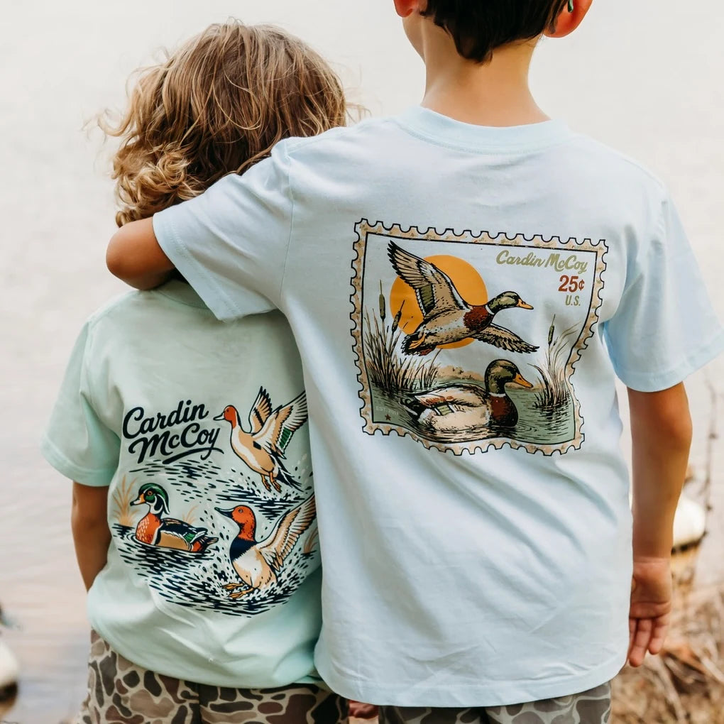 Two boys in pale blue and mint Cardin McCoy duck graphic tees, standing with an arm around each other.