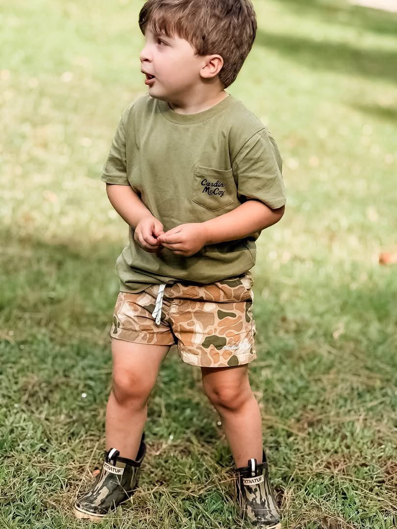 A child wearing an olive Cardin McCoy pocket tee and brown camo shorts outdoors.