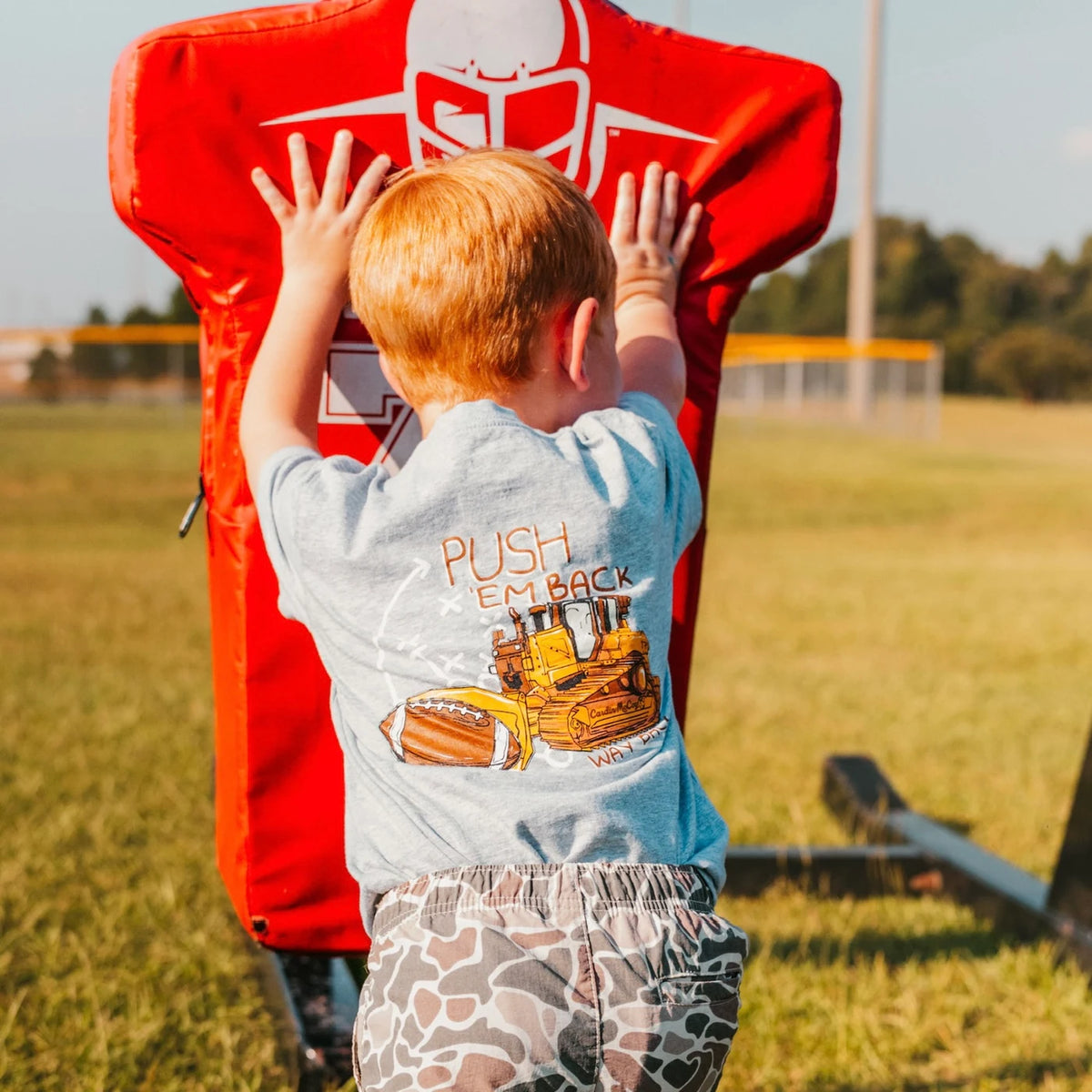 A boy wearing a Cardin McCoy football and bulldozer graphic tee at a football field.